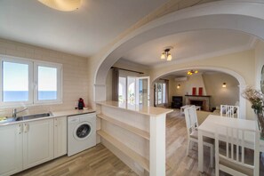 A modern kitchen with a built-in washing machine, a dining area with white furniture, and a fireplace in the background.