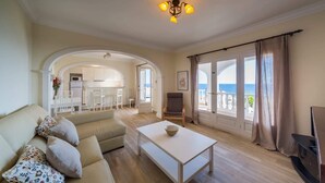 Ein geräumiges Wohnzimmer mit Sofa, Couchtisch und Fernseher. Ein Balkon bietet Blick aufs Meer.