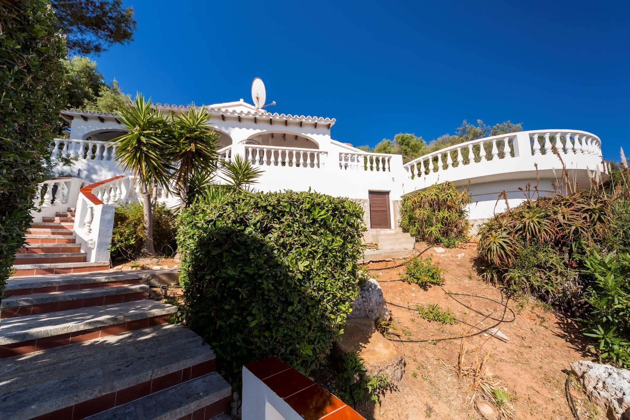 A white villa with a curved balcony, a satellite dish, and a staircase leading to the entrance.