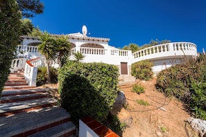 A white villa with a curved balcony, a satellite dish, and a staircase leading to the entrance.