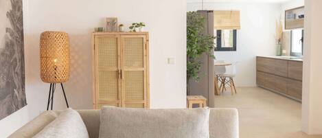 A modern living room with a sofa, a rattan floor lamp, a wooden cabinet with plants, and a view into the kitchen area.
