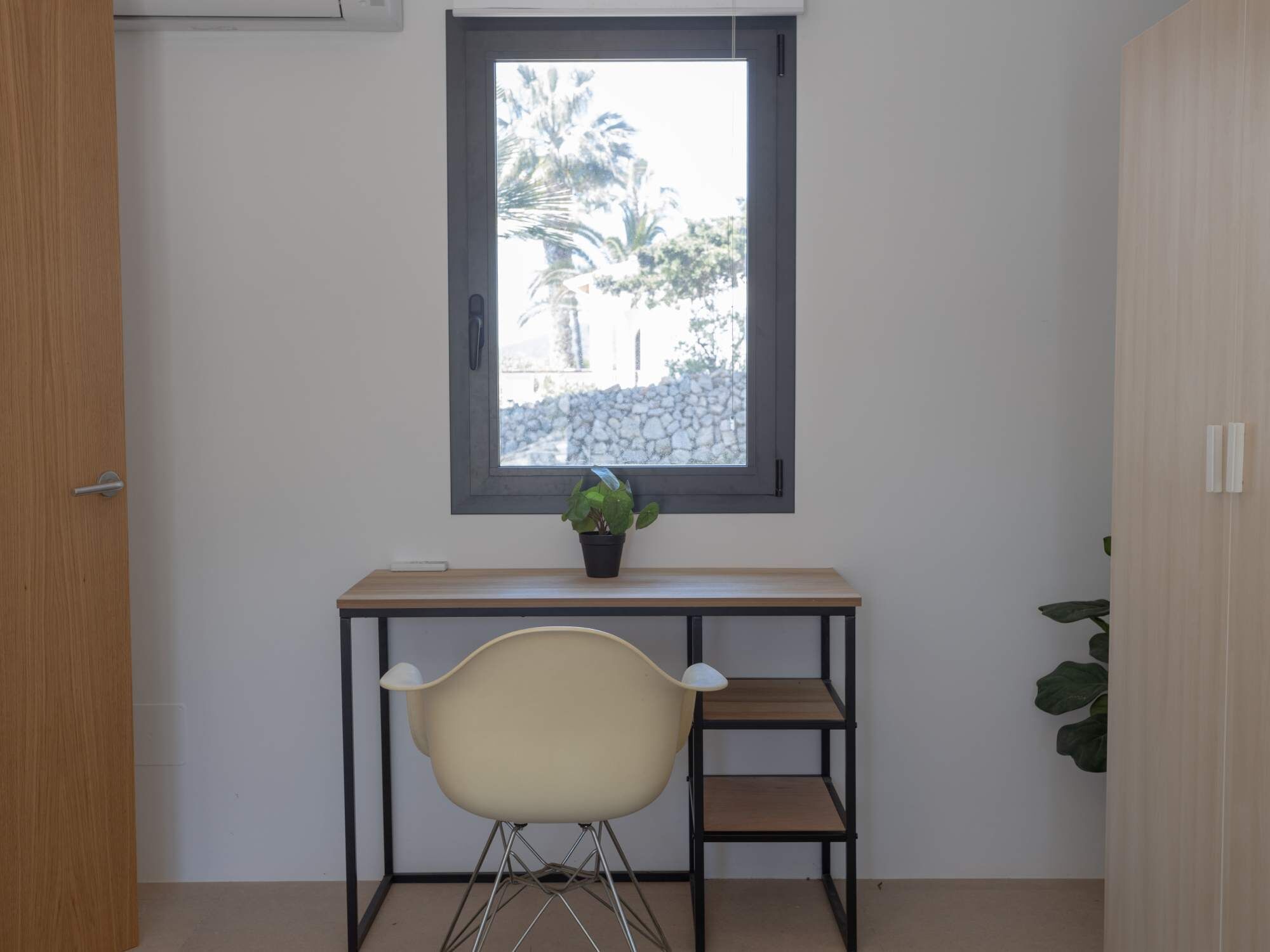 Ein kleines Zimmer mit Fenster, Schreibtisch, Stuhl und einer Topfpflanze.