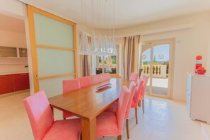 A dining area with a wooden table and pink chairs, a kitchen with red cabinets, and a view of the outdoors through a glass door. - Villa Treya - Star property, stunning views (San Jaime Mediterráneo)