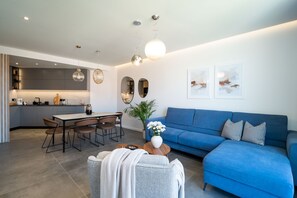 Una sala de estar moderna con un sofá azul, una mesa de comedor con sillas, una mesita de centro y una cocina al fondo.