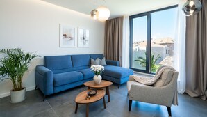 A modern living room with a blue sofa, a beige armchair, a wooden coffee table, and a large window with a view of buildings.