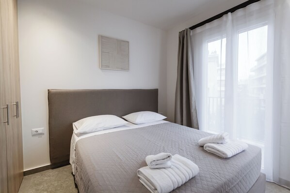 A neatly made bed with white linens and towels, a headboard, a wall-mounted light switch, and a window with sheer curtains.