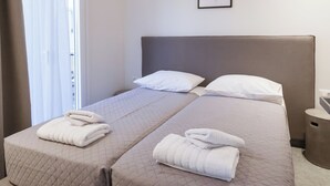 Een tweepersoonsbed met twee kussens en opgevouwen handdoeken, een nachtkastje, een ingelijste foto aan de muur en een raam met gordijnen.