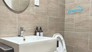 A bathroom with a sink, mirror, and toilet. There are toiletries on the sink and a trash bin next to the toilet.