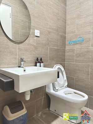 A bathroom with a sink, mirror, and toilet. There are toiletries on the sink and a trash bin next to the toilet. - Ong Kim Wee Residence (Malacca City)