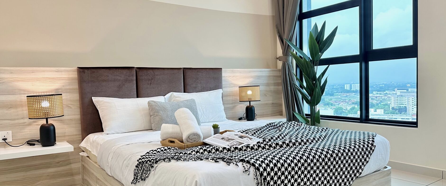 Een moderne hotelkamer met een groot bed, twee bedlampen, uitzicht op de stad door grote ramen en een potplant.