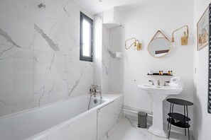 A modern bathroom with a marble wall, a freestanding bathtub, a pedestal sink, and a small black shelf. - Spire View, by Veeve (Paris)