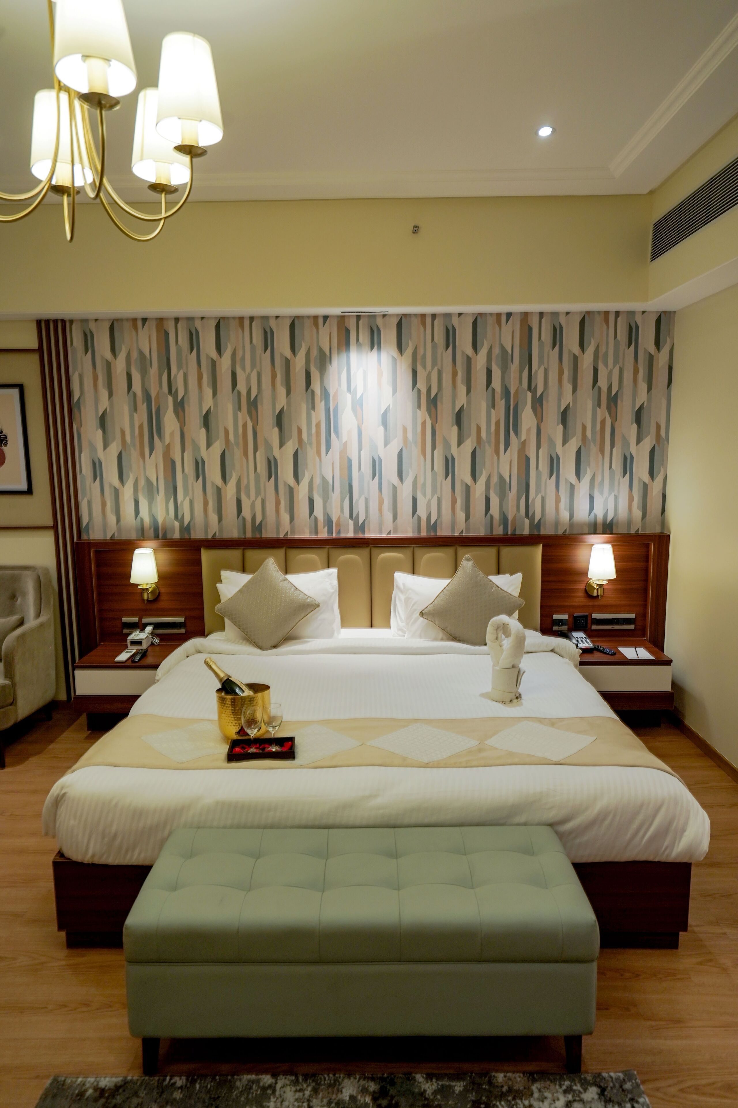 Una habitación de hotel con una cama grande, dos mesitas de noche con lámparas, un cabecero de madera, un papel tapiz estampado y un reposapiés verde.