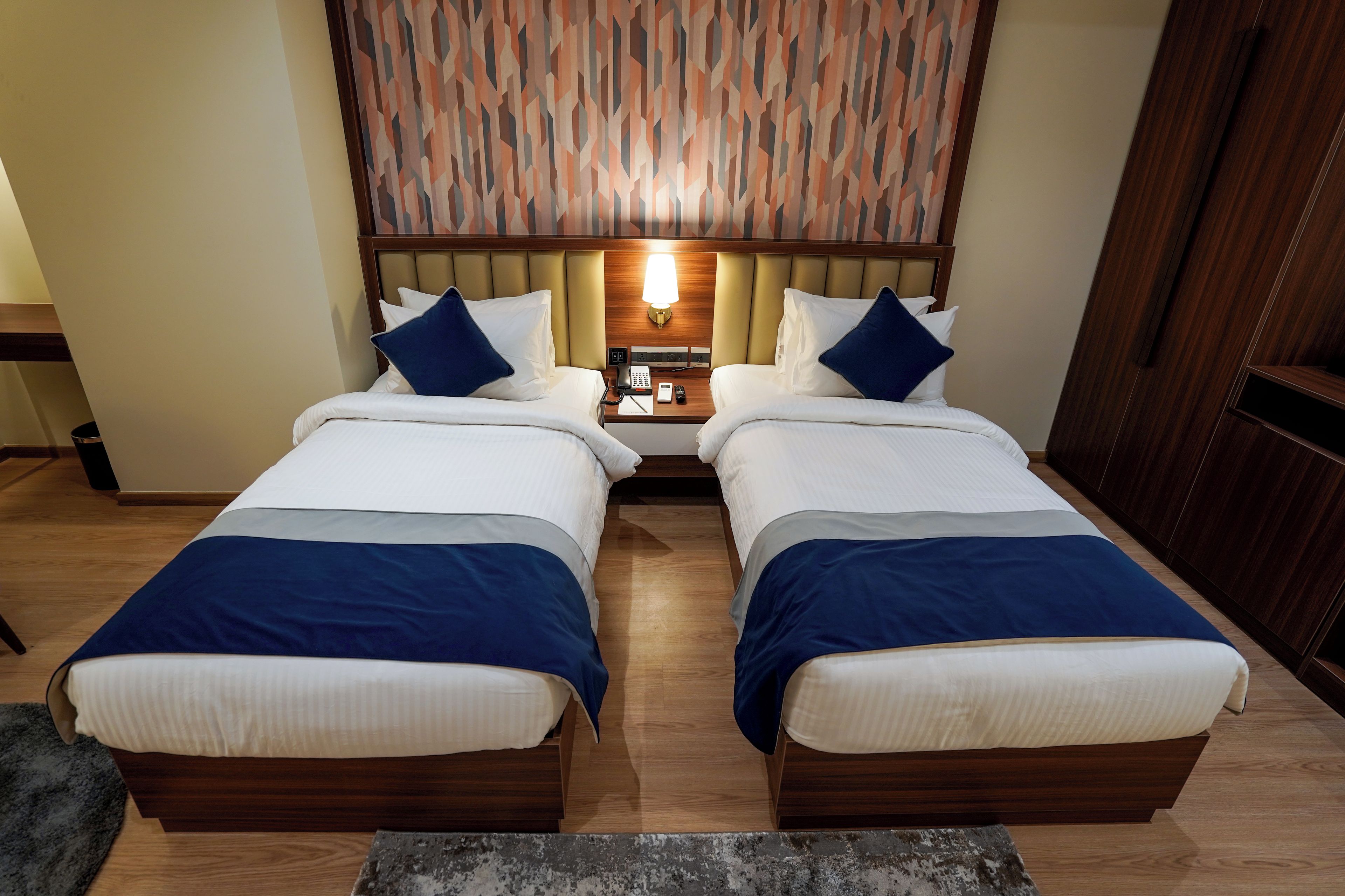 A hotel room with two beds, a nightstand with a lamp, and a patterned wall.