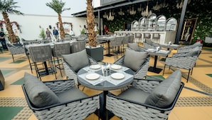 An outdoor dining area with wicker chairs and tables, palm trees, and a bar in the background. - M2 Square by Monday Premium (Nagpur)
