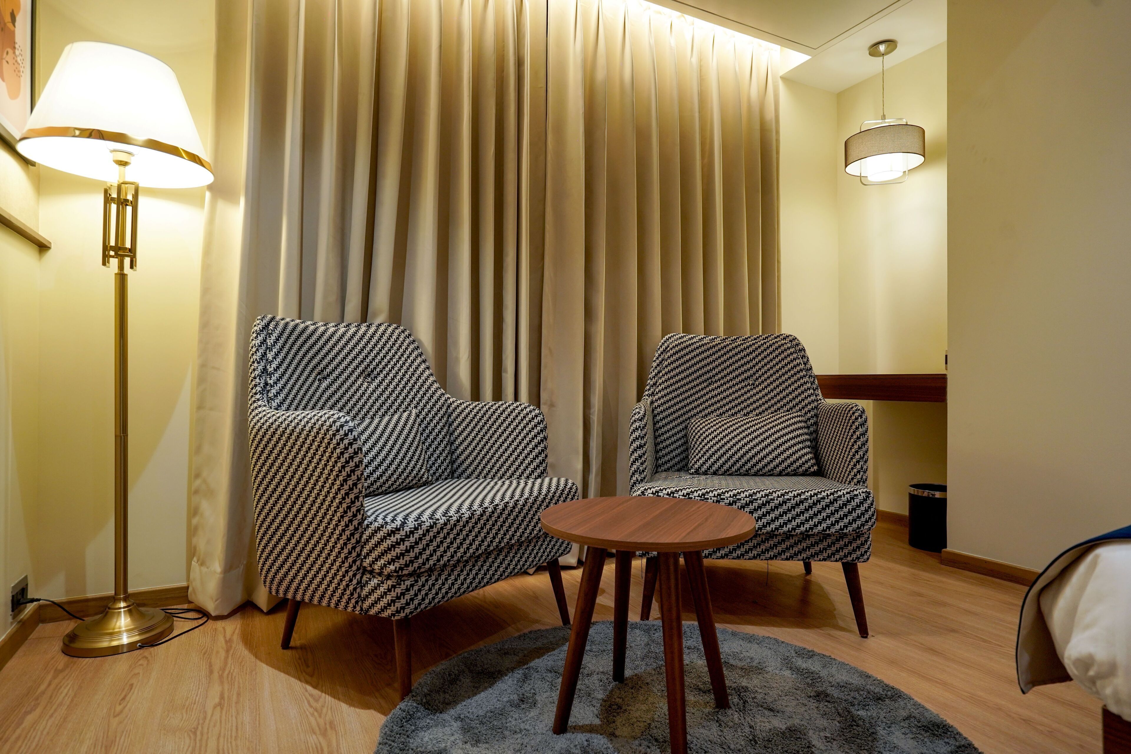 A hotel room with two patterned armchairs, a round wooden table, a floor lamp, and a curtain.