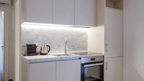 Une cuisine moderne avec des meubles blancs, un plan de travail en marbre, une machine à café, une bouilloire et un four. On peut voir deux petites tables rondes au premier plan.
