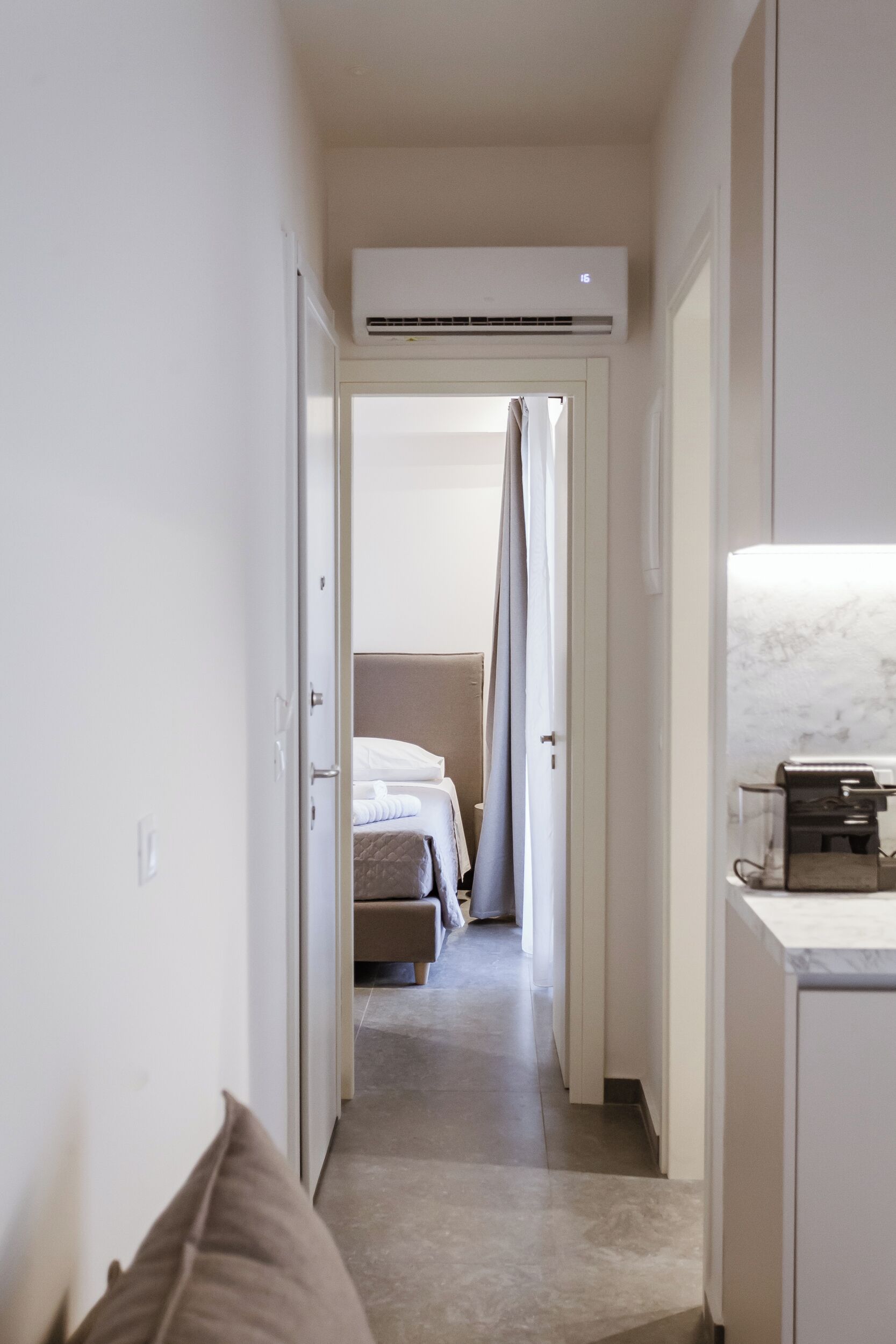 Un couloir étroit, avec un lit au fond, une machine à café sur la droite et une porte sur la gauche.