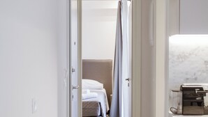 Un couloir étroit, avec un lit au fond, une machine à café sur la droite et une porte sur la gauche.