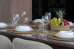 Een eettafel gedekt met witte borden, bestek en heldere wijnglazen, met een geweven mand met fruit en een ananas op tafel.
