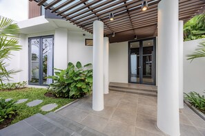 Une entrée extérieure moderne avec un patio couvert, des marches menant à une porte vitrée et une végétation luxuriante.