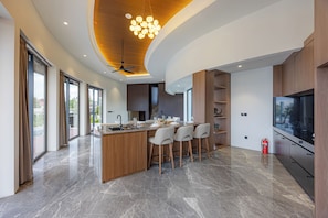 A modern kitchen with a central island, bar stools, a ceiling fan, and a large TV. - Villa Trix Canggu - Elegant 4BR Villa for Families & Friends (Badung)