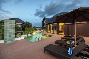 Een zwembad met ligbedden en parasols, omgeven door een houten terras en een gebouw met grote ramen.