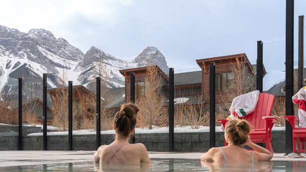 Ein Poolbereich mit zwei Personen, die auf roten Stühlen sitzen, einer Holzhütte und verschneiten Bergen im Hintergrund.
