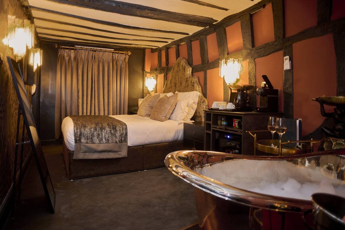Quarto com uma cama grande, cabeceira de madeira, mesa de cabeceira com abajur, pequena mesa com duas taças de vinho e uma banheira cheia de gelo.