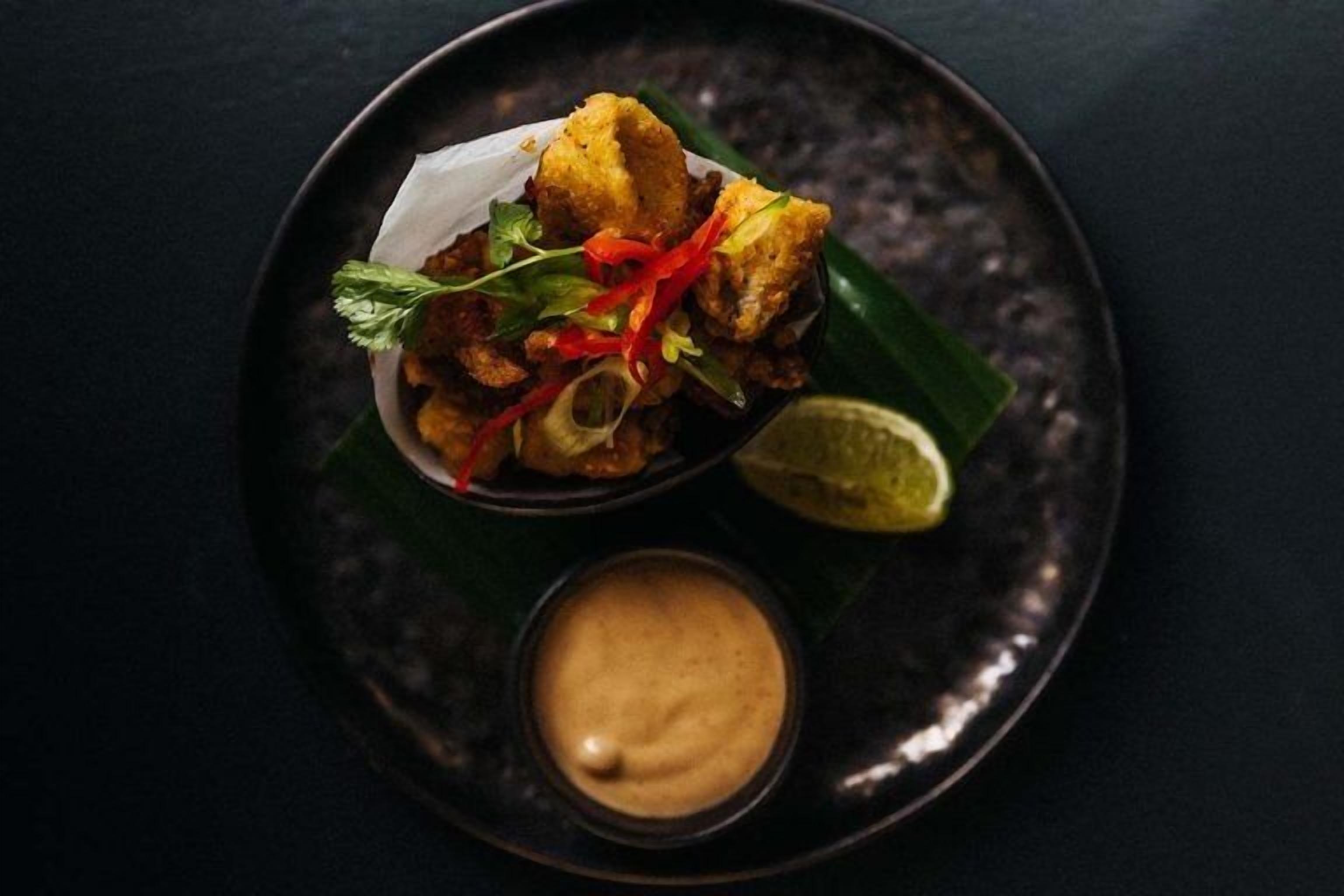 A bowl of fried food with a side of sauce and a lime wedge on a dark plate.