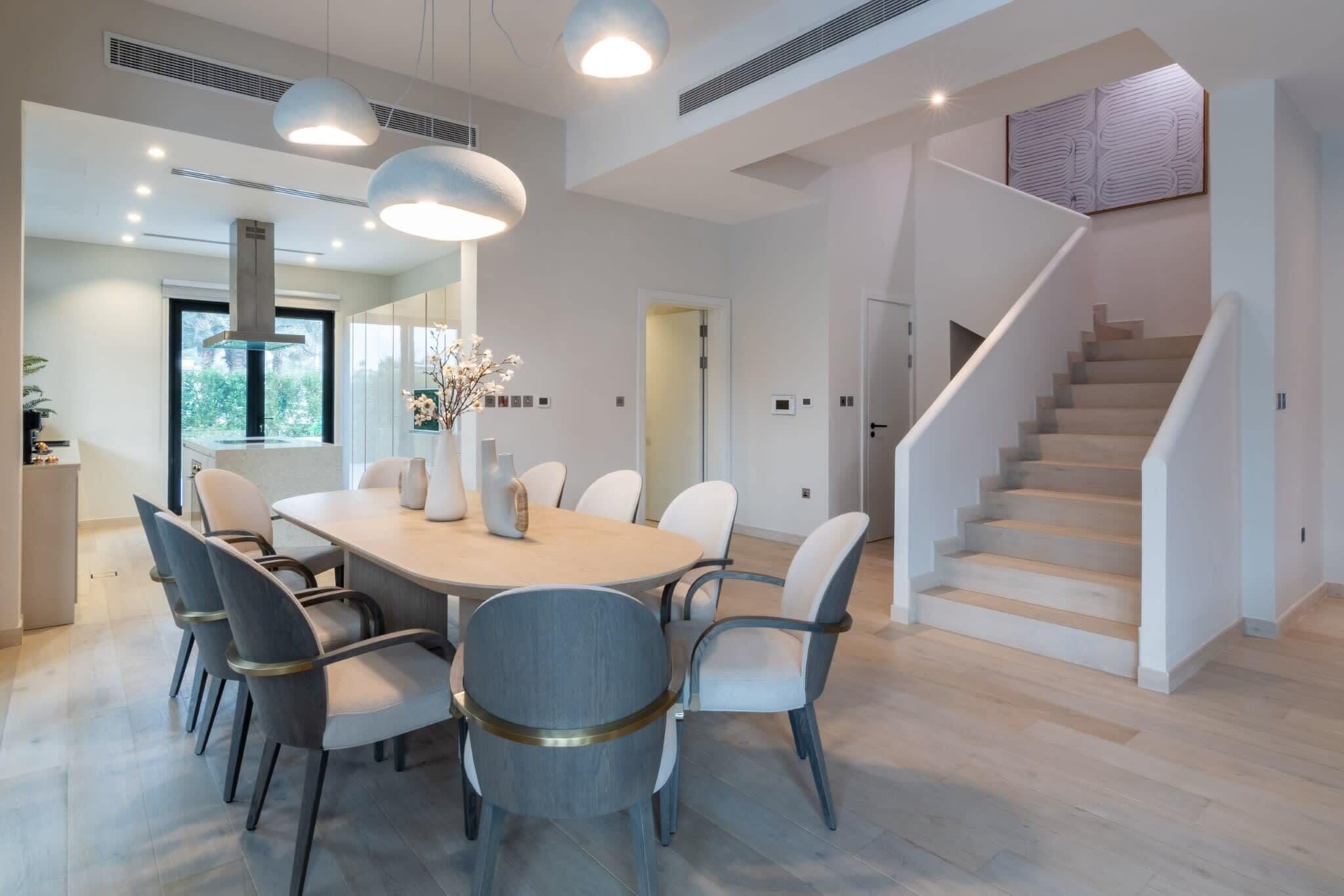 Un espace repas moderne avec une table ronde, plusieurs chaises, des luminaires suspendus et un escalier.