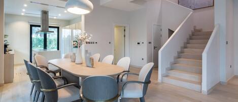 Un espace repas moderne avec une table ronde, plusieurs chaises, des luminaires suspendus et un escalier.