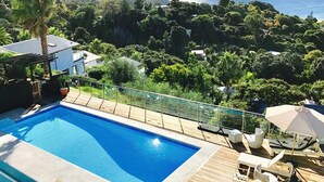 A swimming pool with a wooden deck, lounge chairs, and a glass railing overlooking a forested area.