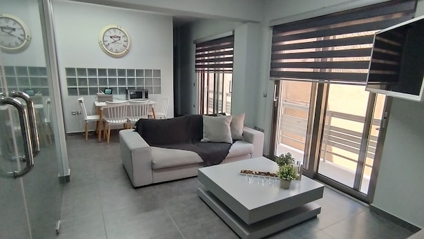 Una sala moderna con un sofá, una mesa de centro y un comedor con una mesa y sillas. Hay relojes en la pared y un televisor montado allí.