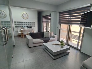 A modern living room with a sofa, coffee table, and a dining area with a table and chairs. There are clocks on the wall and a TV mounted on the wall. - Academys Modern Apartment by Aesthetic & Luxurious (Athens)