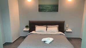 Una cama bien hecha con edredón gris, dos almohadas y una toalla doblada. Hay dos mesitas de noche con lámparas y una pequeña planta en maceta sobre una de ellas.