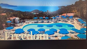 Un resort con una piscina grande rodeada de sombrillas azules y sillas de descanso, con vistas a una zona costera con montañas y cielo despejado.