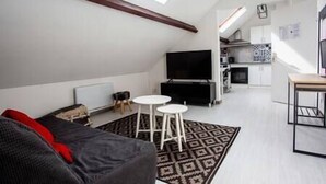 A modern loft apartment with a high ceiling, a black sofa, a TV, and a small kitchen area.