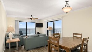 A living room with a sofa, a dining table with chairs, a ceiling fan, and a TV mounted on the wall. - Two Bedroom Beachfront End Unit Condo with Pool. (Ocean City)