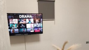 A room with a television displaying a drama channel, a small table with a vase, and a blue carpet. - Ermou Junior Suite by Aesthetic & Luxurious Living (Athens)