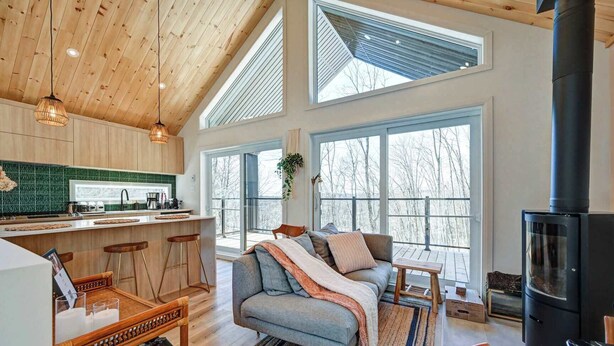 A spacious living area with a large sofa, a coffee table, and a fireplace. There is a kitchen area with a bar and stools. The room has large windows and a high ceiling with wooden beams.