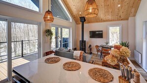 A spacious living area with a large wooden ceiling, a central island with woven placemats, a built-in wood stove, and a large window overlooking a forested area.