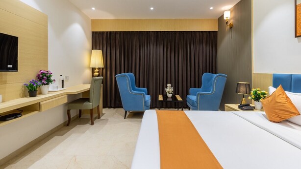 A hotel room with a bed, two blue armchairs, a desk with a lamp, a TV, and a large window with curtains. - JANKI PALACE (Bhopal)
