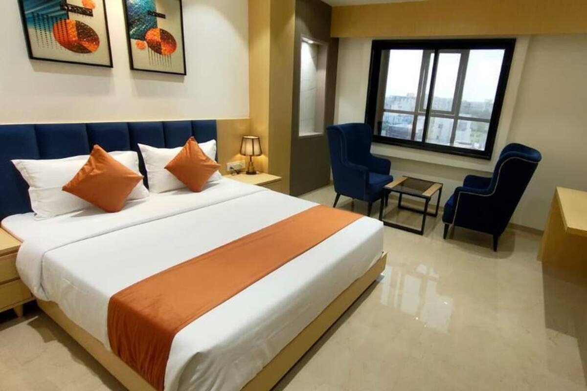 Quarto de hotel com uma cama grande, duas poltronas azuis, uma mesa pequena e uma janela com vista para a cidade.
