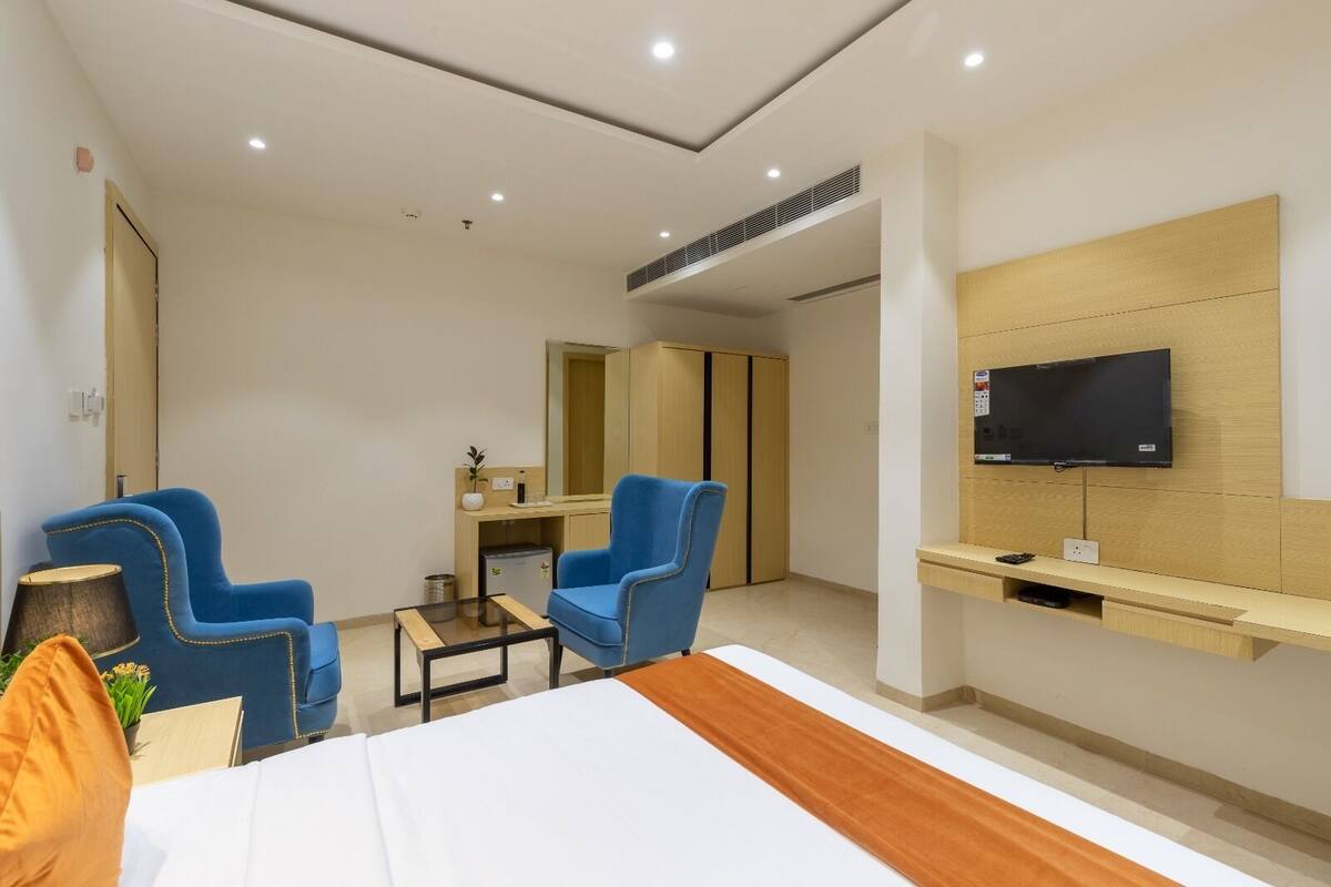 Quarto de hotel com uma cama, duas poltronas azuis, uma mesa pequena, televisão presa à parede e um armário de madeira.
