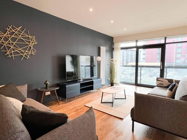 A modern living room with a large flat-screen TV, dark walls, and a large window offering a city view.