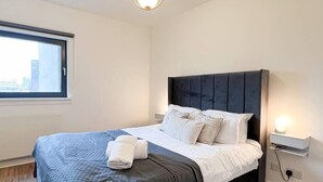 A modern hotel room with a large bed, a window with a city view, and a bedside table with a lamp. - Lovely Spacious Apartment with Hydro & SECC (Glasgow City)