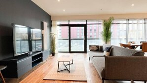 A modern living room with a flat-screen TV, a sofa, a coffee table, and a large window offering a city view. - Lovely Spacious Apartment with Hydro & SECC (Glasgow City)