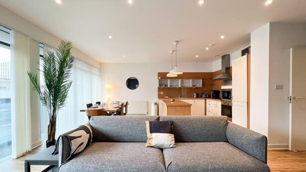 A modern living room with a grey sofa, a dining table, and a kitchen area. - Lovely Spacious Apartment with Hydro & SECC (Glasgow City)