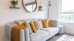 Ett modernt vardagsrum med en grå soffa, en senapsgul pläd och mönstrade kuddar. Det finns två väggmonterade blomkrukor och en golvlampa.