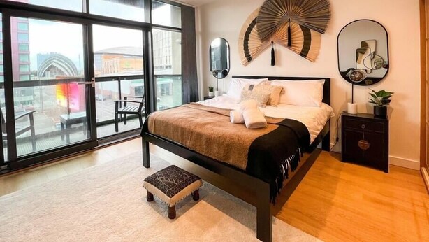 Un dormitorio moderno con un ventanal grande, una cama con una manta color beige, un taburete pequeño, una mesita de noche con una planta y un ventilador de pared.